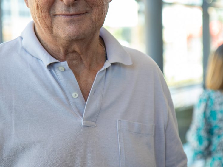 Image of An older man wearing glasses and a light purple polo shirt holds a plate of dessert, looking slightly to the side. The background is softly blurred, with another person in a floral dress visible.