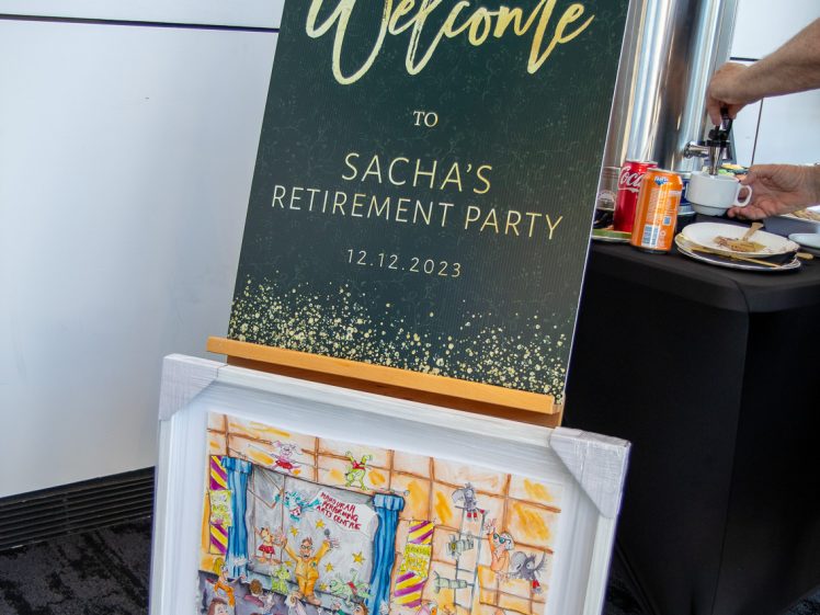 Image of A sign reading Welcome to Sacha’s Retirement Party 12/12/2023 stands on an easel, with a colourful framed caricature below. A person’s hand is seen at a table with drinks and snacks in the background.