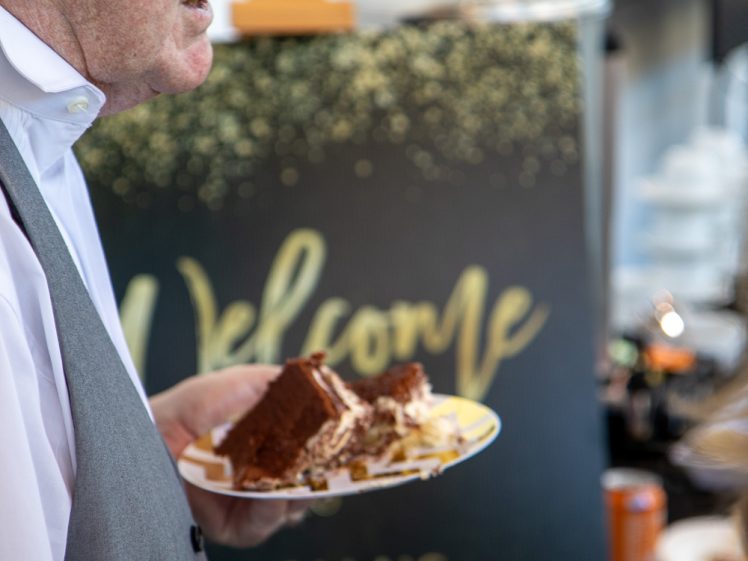 Image of A man in a white shirt and grey waistcoat holds a plate with a slice of chocolate cake. A blurred sign in the background reads, “Welcome SACHA’S RETIREMENT PARTY.”.