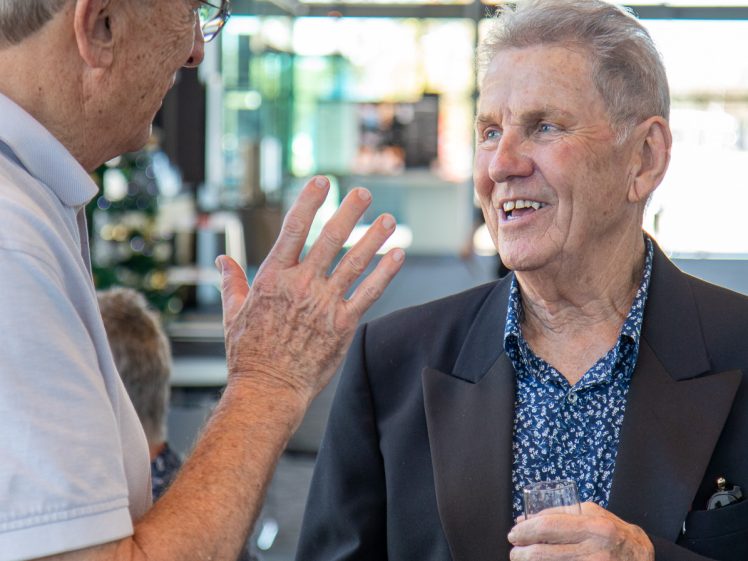 Image of Two older men are having a lively conversation indoors. One man is gesturing with his hand whilst the other, dressed in a blazer and holding a wine glass, smiles warmly. The background is bright and softly blurred.