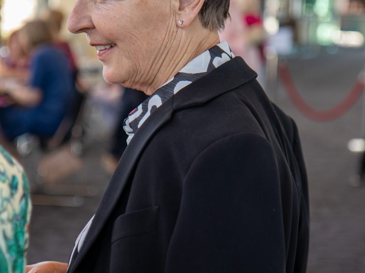 Image of A woman with short brown hair and a black blazer smiles whilst standing indoors, with blurred people and glass windows in the background.
