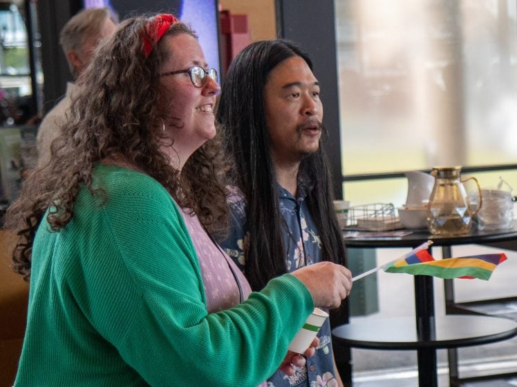 Image of Two people stand indoors, smiling and holding a small rainbow flag. One has long curly hair, glasses, and a green cardigan; the other has long straight hair and a floral shirt. There are tables and dishes in the background.