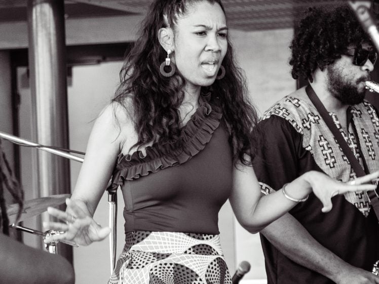 Image of A woman sings passionately into a microphone on stage while a man beside her plays the saxophone. Both are wearing patterned clothing, and musical instruments are visible in the background. The image is in black and white.