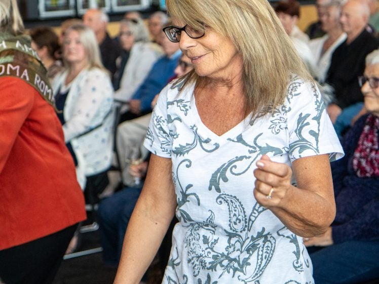 Image of A woman with long blonde hair and glasses, wearing a white shirt with a grey floral pattern, is dancing and smiling at a crowded indoor event with seated people in the background.