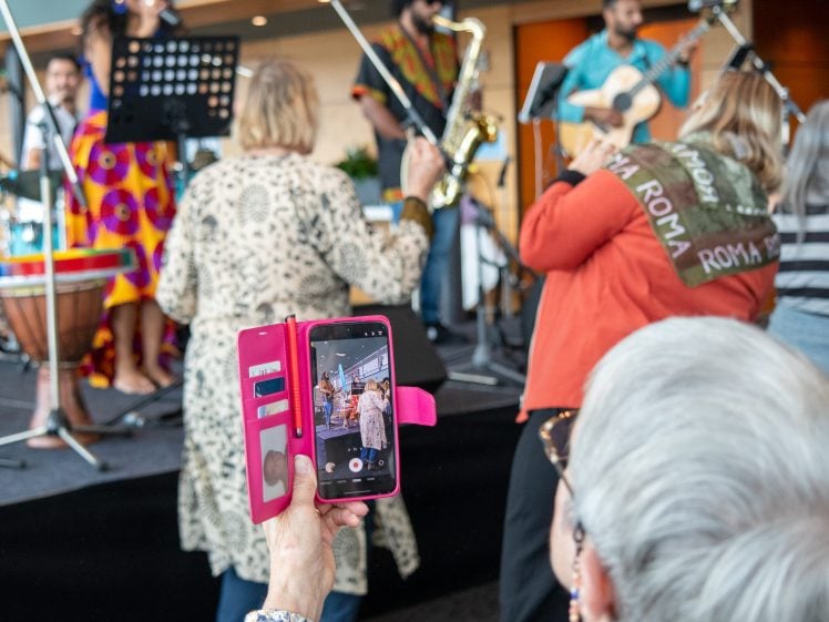 Image of A woman holds up a pink mobile to record a lively indoor musical performance, with musicians singing and playing instruments on stage and audience members dancing and enjoying the event.