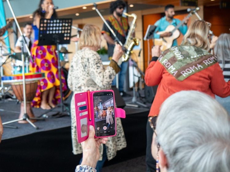 Image of A group of musicians perform on stage as people dance nearby. In the foreground, a person holds a pink phone case, recording the scene, whilst others enjoy the lively atmosphere.