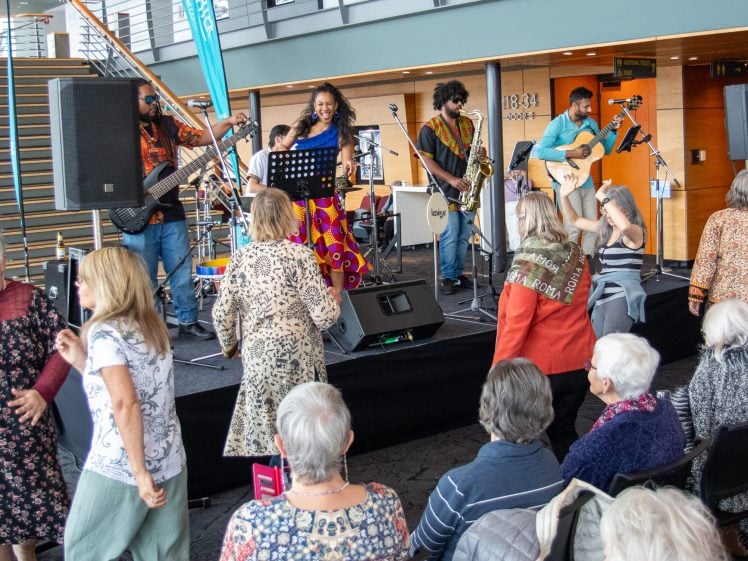 Image of A band performs on an indoor stage while older adults, some seated and some dancing, enjoy the music. The setting appears lively, with bright lighting and modern architecture in the background.