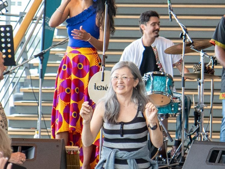 Image of A woman in a colourful skirt sings on stage with a live band, while a smiling woman in a striped shirt dances in the foreground near the audience. Stairs and musical instruments are visible in the background.