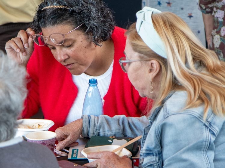 Image of Two women sit closely at a table, concentrating on something in front of them. One wears glasses and a red jumper, the other has blonde hair and a headband. Bowls, cups, and a water bottle are on the table.