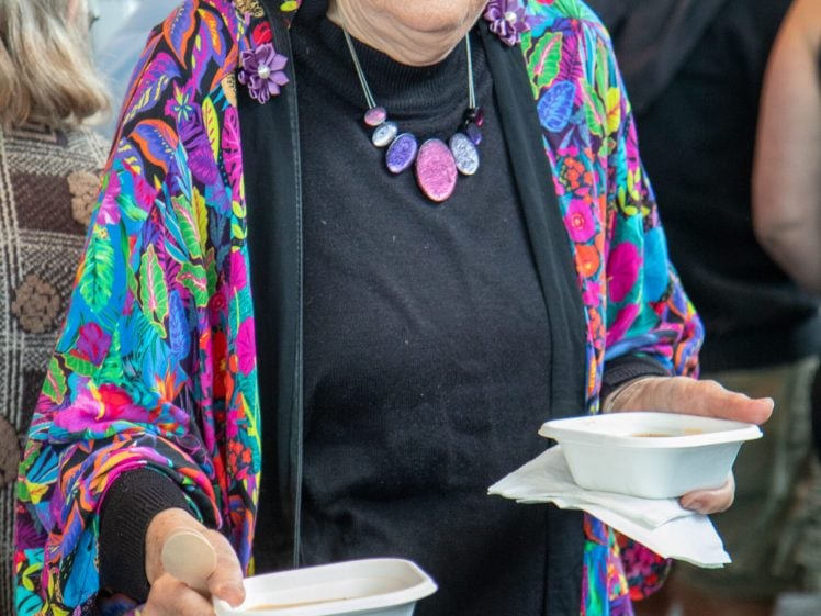 Image of An older woman wearing a colourful floral jacket, black hat, and purple glasses holds two white food containers in her hands whilst standing among a crowd.