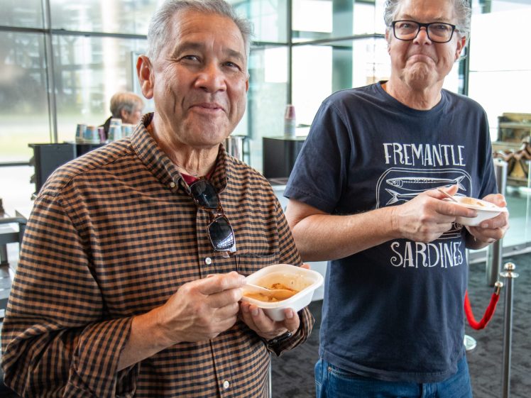 Image of Two older men standing indoors, smiling while holding bowls of food and spoons. One wears a checked shirt and sunglasses, the other a dark graphic t-shirt and glasses. A modern, glass-walled background is visible.