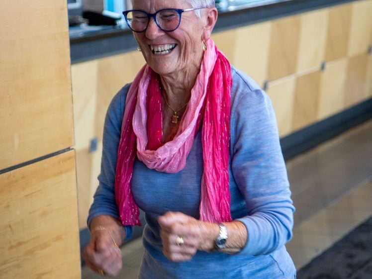 Image of An older woman with short grey hair, glasses, and a pink scarf smiles joyfully indoors, clenching her fists in excitement. She wears a blue shirt and jeans, standing near a wooden wall and a counter in a bright space.