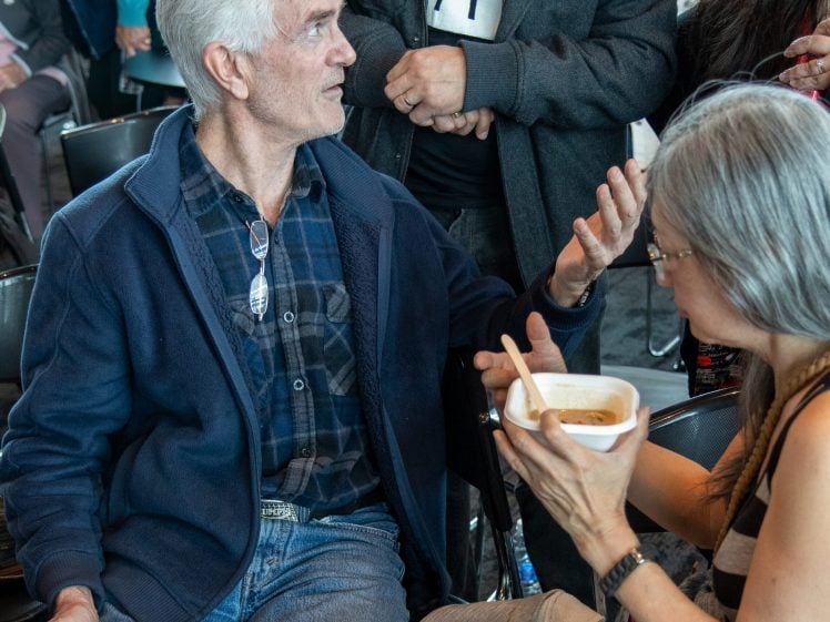 Image of An older man sitting and holding a bowl of soup talks animatedly with a woman beside him who is eating. Several people stand and sit nearby, creating a lively atmosphere.