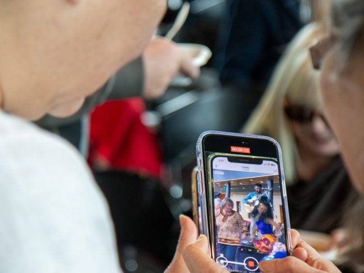 Image of A person holds a mobile phone and records a video of a small group of people performing or celebrating indoors, while another person looks on from the side. The phone screen shows the scene being recorded.
