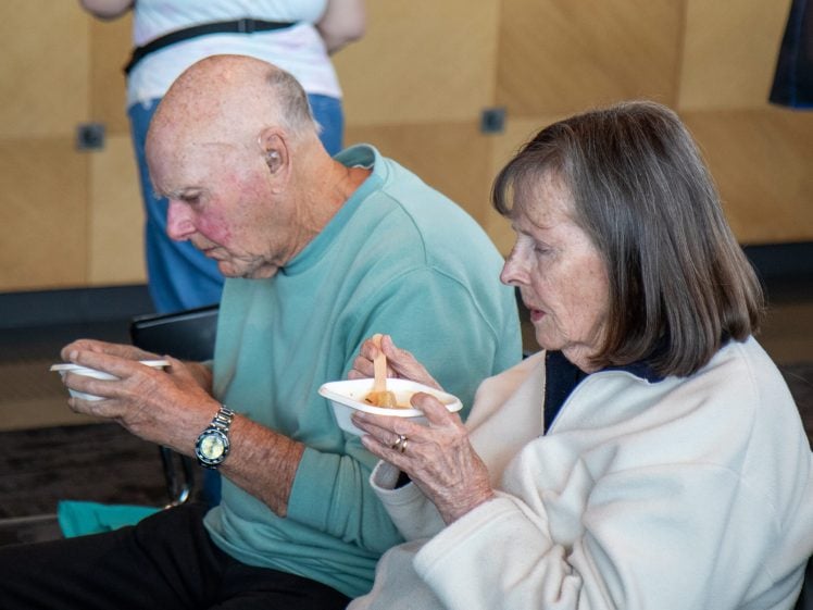 Image of An older man and woman sit side by side, eating from small white bowls with wooden spoons. They appear focused on their food. The background is indoors with wooden panels on the wall.
