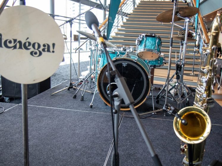 Image of Close-up of a stage set-up with a drum kit, saxophone, microphones, and a percussion instrument labelled tenéga! in the foreground. Wooden stairs and bright windows are visible in the background.