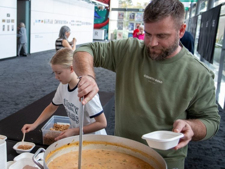 Image of A man ladles soup from a large pot into a bowl while a young girl beside him serves herself toppings at an indoor event with artwork displayed on the walls in the background.