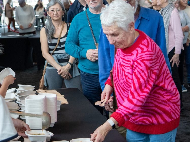Image of A group of people stands in a queue at a buffet, with an elderly woman in a pink and red jumper reaching for a bowl of food whilst others wait behind her. A server ladles food into bowls on a table.