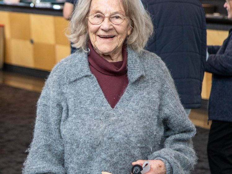 Image of An elderly woman with grey hair and glasses smiles while holding a walking stick. She is wearing a maroon polo neck under a grey jumper, standing indoors with people in the background.