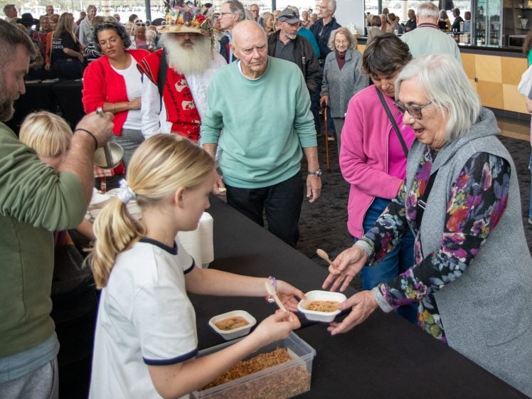 Image of A young girl serves food to an elderly woman at a crowded indoor event. People of various ages wait in a queue, and there is a container of cereal and bowls on the table. Everyone appears engaged and lively.
