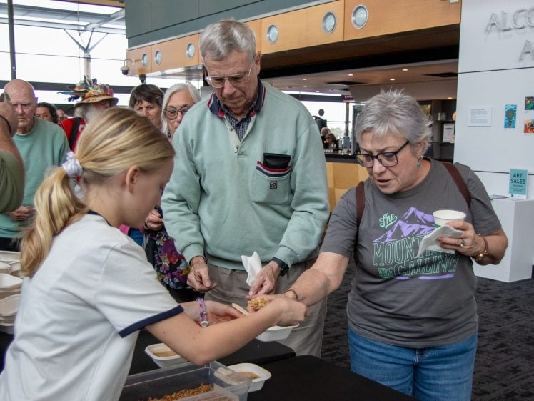 Image of A young girl serves granola into the hand of an older woman at a food table, while an older man stands beside them. Several people queue in line in a modern indoor setting.