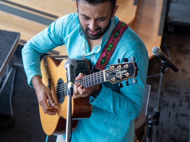 Image of A man in a light blue shirt plays an acoustic guitar and sings into a microphone. He has a red patterned strap on his guitar and is performing indoors with musical equipment around him.