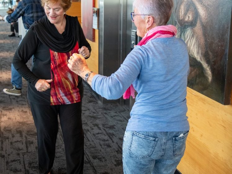 Image of Two older women smiling and dancing together in a corridor, with a large portrait of a man visible on the wall behind them. One woman wears a colourful top, and the other a blue shirt and pink scarf.