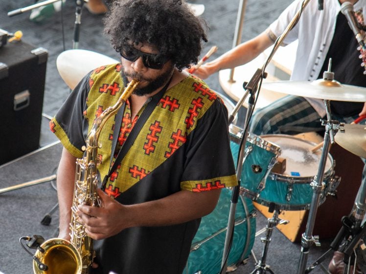 Image of A musician wearing sunglasses and a black shirt with colourful patterns plays the saxophone on stage, whilst another person behind him plays the drums. Microphones and musical equipment are visible.