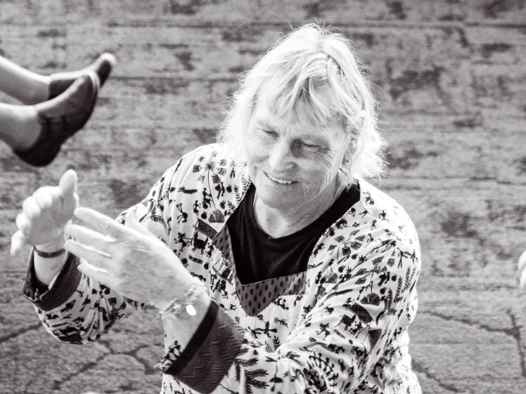 Image of An older person with fair hair smiles while raising their hands, wearing a patterned jumper. Another person's legs are visible in the top left, suggesting playful interaction. The image is black and white.