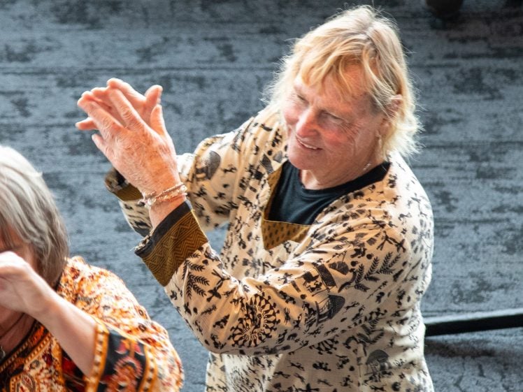 Image of Two older adults, one in the foreground and one in the background, wearing patterned shirts. The person in the background is smiling and clapping, while both are engaged in an indoor group activity.