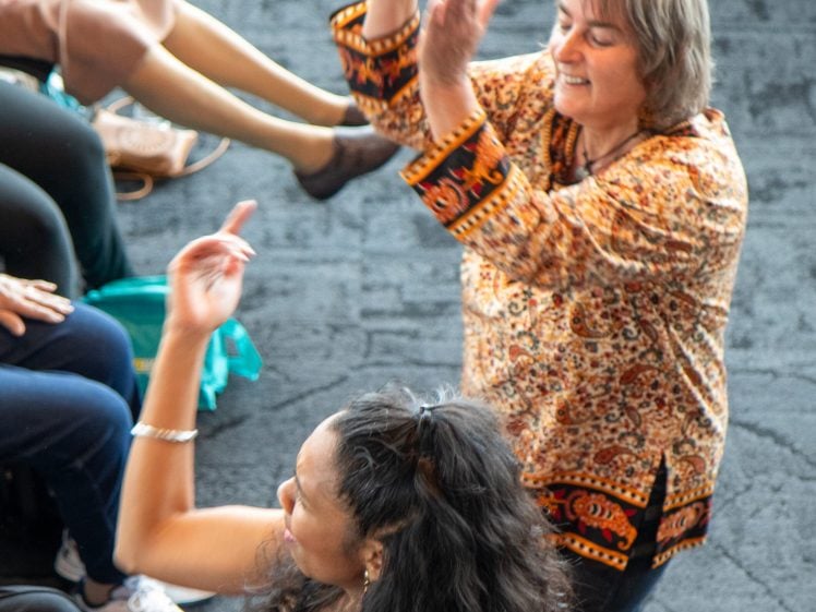 Image of Two women joyfully dance with raised arms among a seated group. One wears a colourful patterned top and the other a blue dress. The background shows other people clapping and smiling.