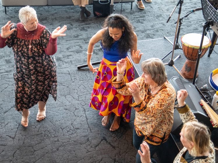 Image of Four women stand barefoot in a circle, dancing joyfully indoors. They wear colourful dresses and tops, raising their arms in the air, with musical instruments visible nearby. The atmosphere is lively and celebratory.
