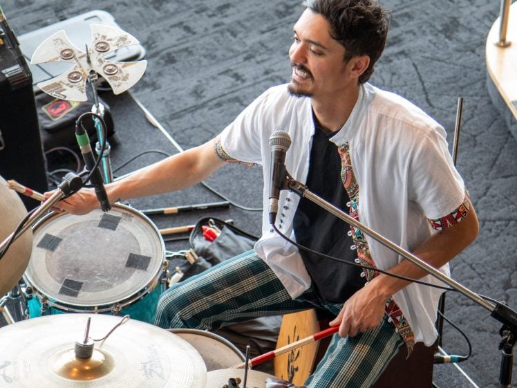 Image of A smiling musician wearing a white shirt and patterned trousers plays drums and percussion instruments, surrounded by microphones and music equipment.