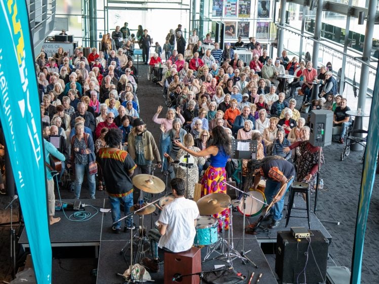 Image of A band performs on stage in front of a large seated audience in a bright indoor venue with floor-to-ceiling windows. The crowd watches attentively, filling the space with lively energy.