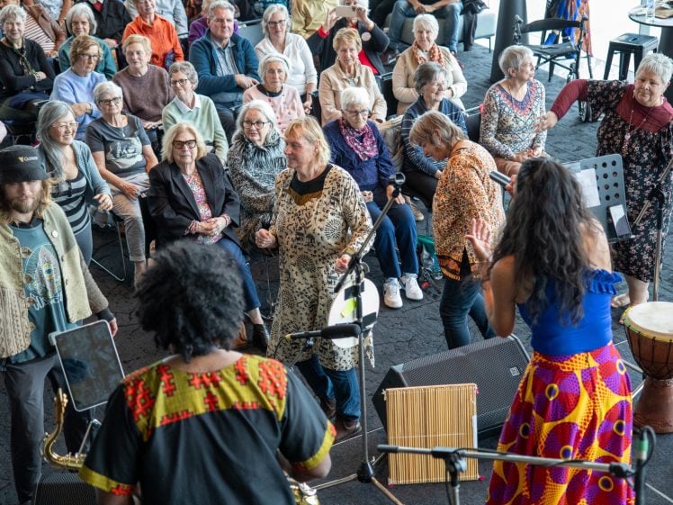 Image of A diverse group of older adults, some seated and some dancing, enjoy a lively indoor musical performance by musicians in colourful clothing, with drums and other instruments visible. The audience looks engaged and joyful.