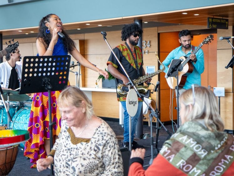 Image of A lively band performs indoors as a woman sings and others play instruments. Two older women in the foreground dance and enjoy the music. The atmosphere is joyful and colourful.