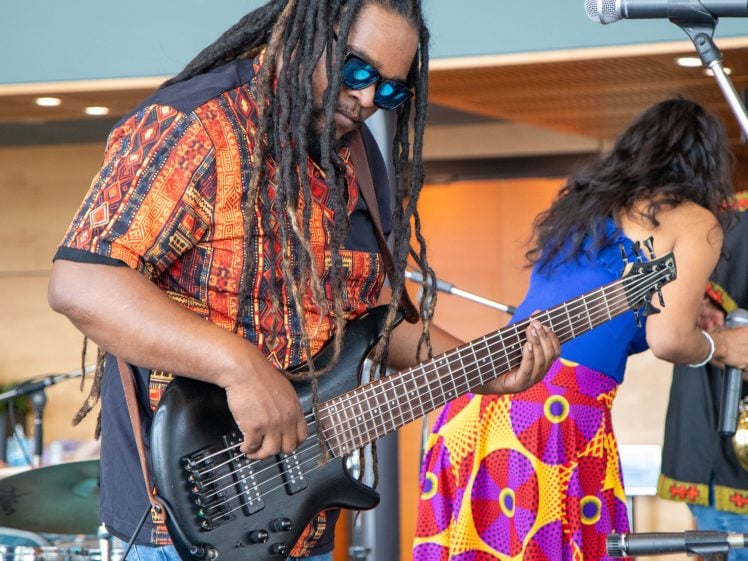 Image of A musician with long dreadlocks and sunglasses plays a black electric bass guitar on stage. He wears a colourful patterned shirt. A woman in a vibrant skirt with geometric designs stands nearby. Instruments and microphones are visible.