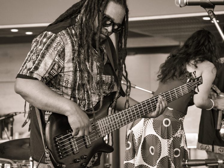 Image of A musician with long dreadlocks and sunglasses plays a bass guitar on stage, whilst another performer stands nearby. The scene is in black and white, capturing a lively musical performance.