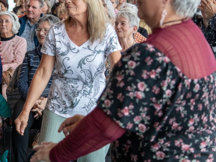 Image of Two older women stand and move to music in a crowded indoor venue, while an audience of seated people watches and smiles. The environment appears lively and cheerful.