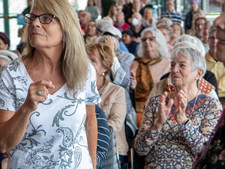 Image of A woman with glasses stands and gestures in a crowded room full of older adults, some seated and applauding, while others look on attentively. The atmosphere appears engaged and lively.