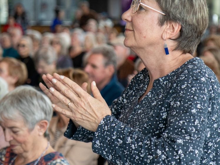 Image of An older woman with short grey hair and glasses stands and applauds in a crowded indoor setting, with several people seated and watching attentively in the background.