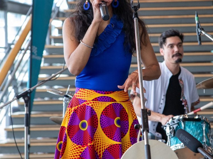 Image of A woman sings into a microphone on stage, wearing a bright blue top and a colourful patterned skirt. A drummer plays behind her. The drum has the word Ichéga! printed on it. They are performing indoors near a staircase.