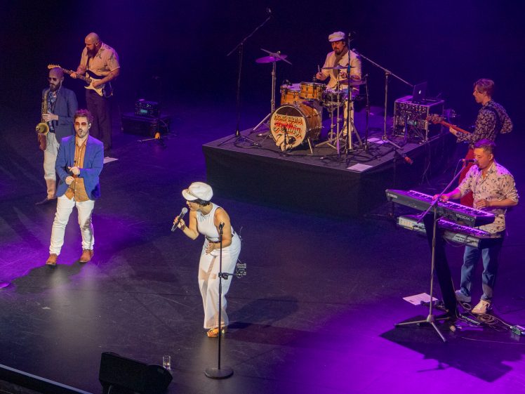 Image of A six-member band performs on stage under purple lights. The members play saxophone, trumpet, drums, guitar, and keyboard, whilst one person sings into a microphone at the front of the stage.