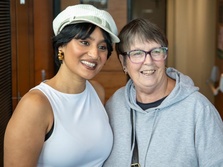 Image of Two women smiling and posing together indoors. One wears a white sleeveless top and a white hat, the other wears glasses and a grey hoodie. They appear happy and are standing close to each other.