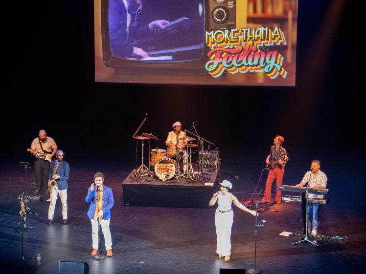 Image of A band performs on stage with singers, saxophonist, guitarist, drummer, and keyboard player. Above them, a screen shows a vintage TV with a pianist and the text More Than a Feeling in colourful letters.