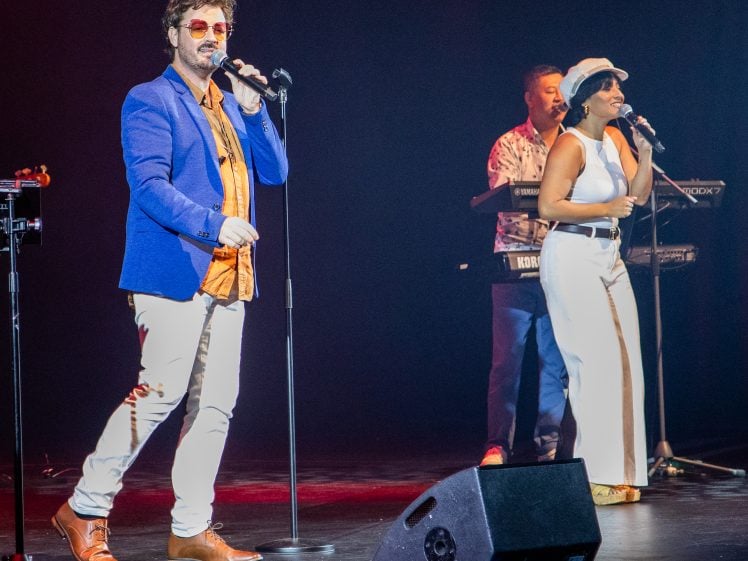 Image of A man in a blue blazer and sunglasses sings into a microphone on stage beside a woman in white trousers and a hat, with a keyboard player performing in the background under stage lights.