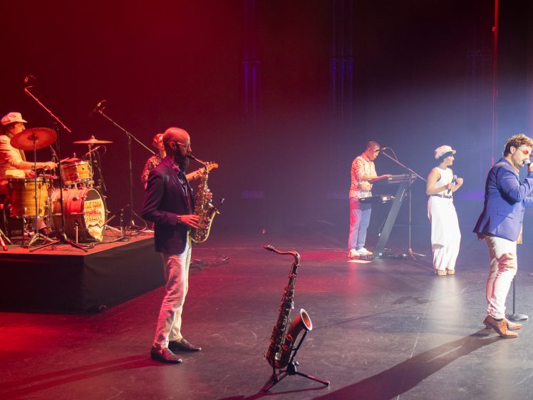 Image of A band performs on stage under colourful lighting. A drummer, saxophonist, keyboard player, female vocalist, and male singer are mid-performance, with instruments and microphones set up around them.