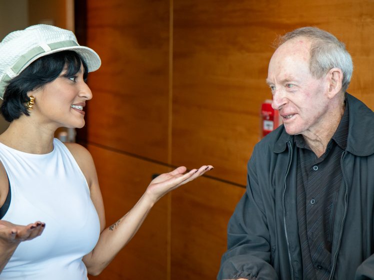Image of A woman in a white hat and sleeveless top gestures while talking to an older man in a dark jacket. They are indoors, standing in front of a wooden wall, and both appear to be engaged in conversation.