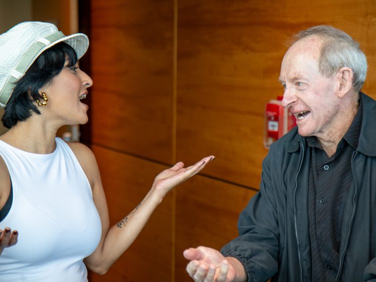 Image of A woman in a white hat and sleeveless top animatedly talks with an older man in a black jacket. Both are smiling and gesturing with their hands in front of a wooden wall with a fire alarm in the background.