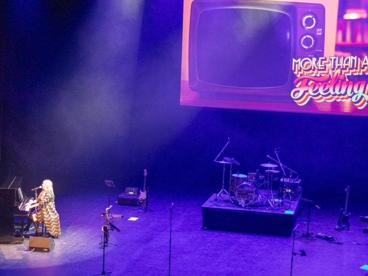 Image of A musician plays piano and sings on stage under a spotlight, with empty instruments nearby. A large screen above displays an old TV and the words More Than A Feeling in colourful text.